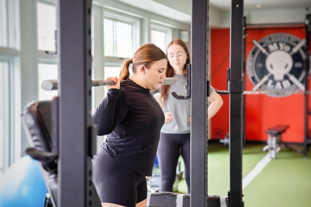 woman doing squats with an empty weight bar with a MYo Lab trainer in the back in the MYo lab gym