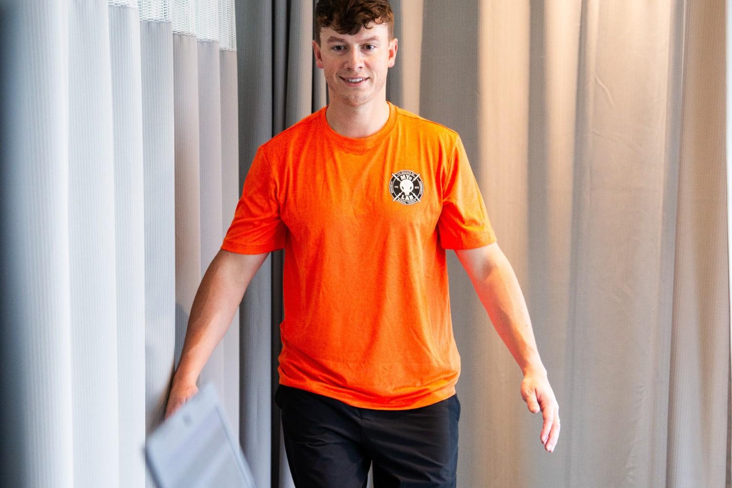 A man standing indoors wearing a orange t-shirt. He has short sleeves and is positioned against a wall.