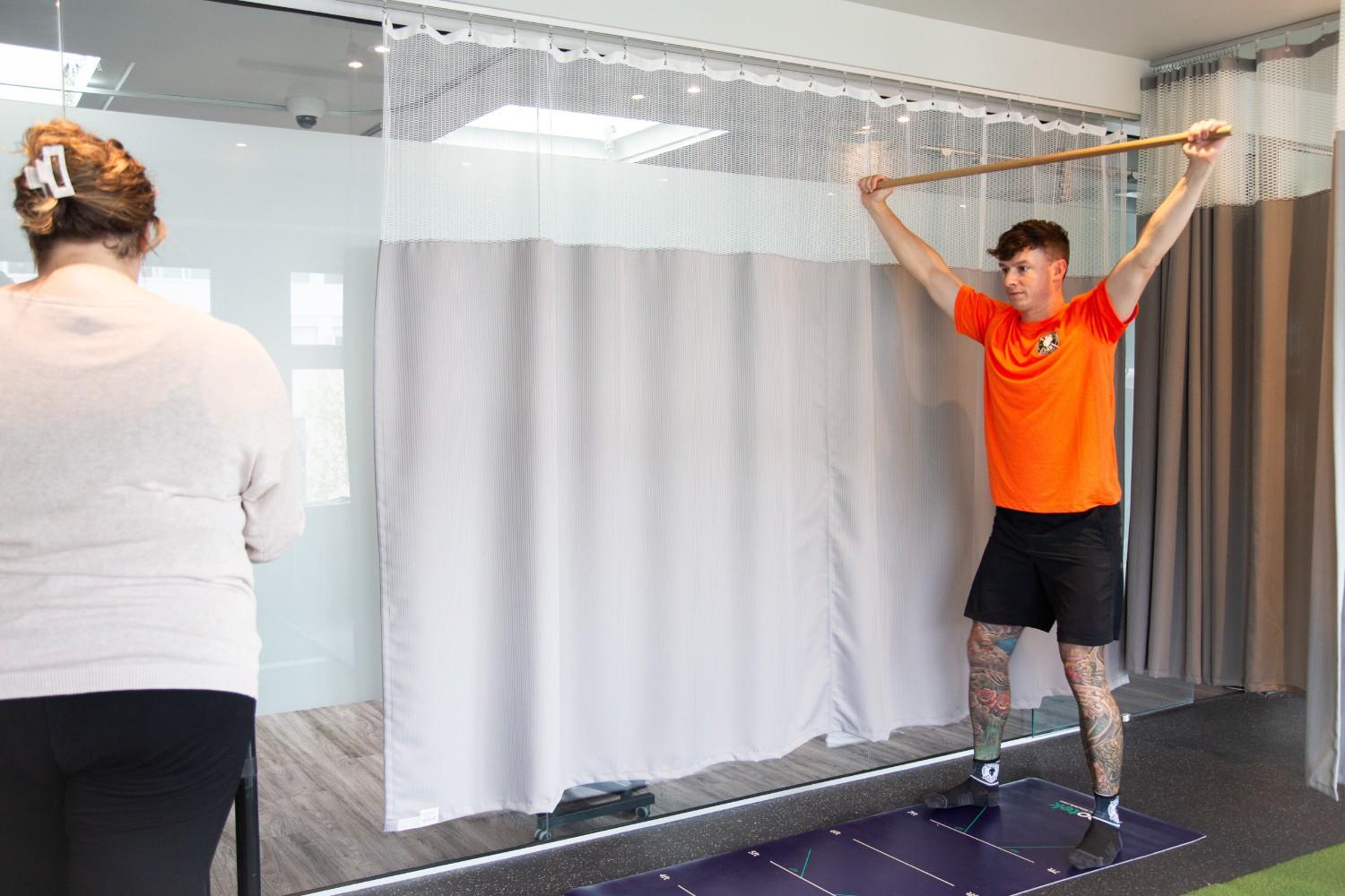 A man in an orange shirt holds a wooden stick overhead while standing on a mat.