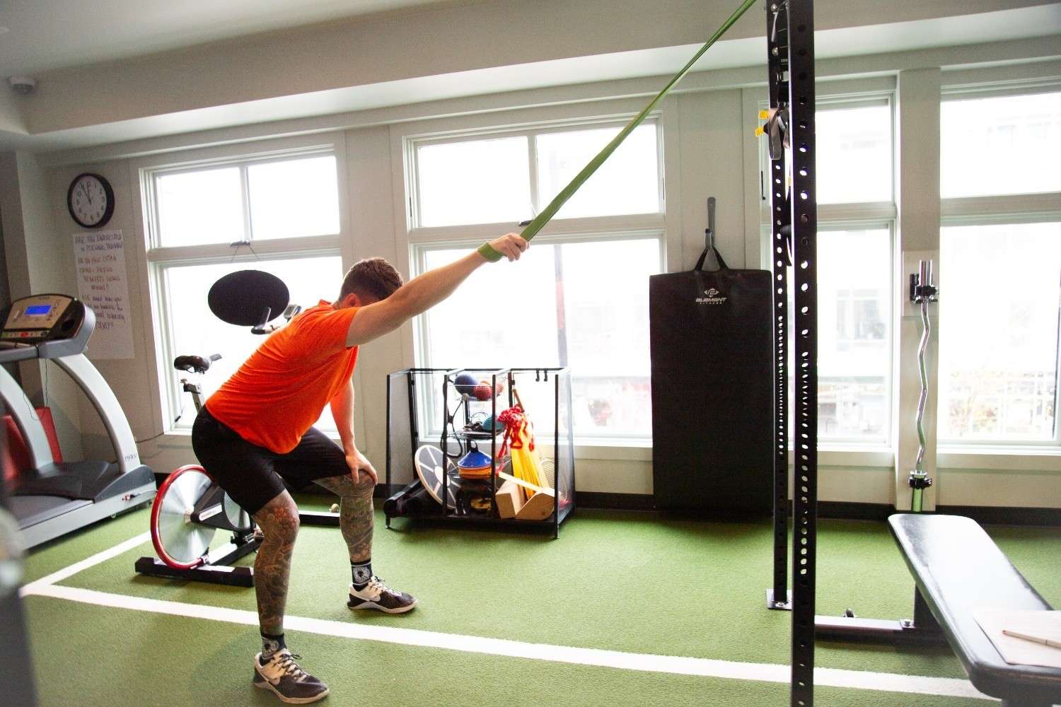 A man wearing an orange shirt and black shorts performs an exercise with a resistance band in a bright gym space.