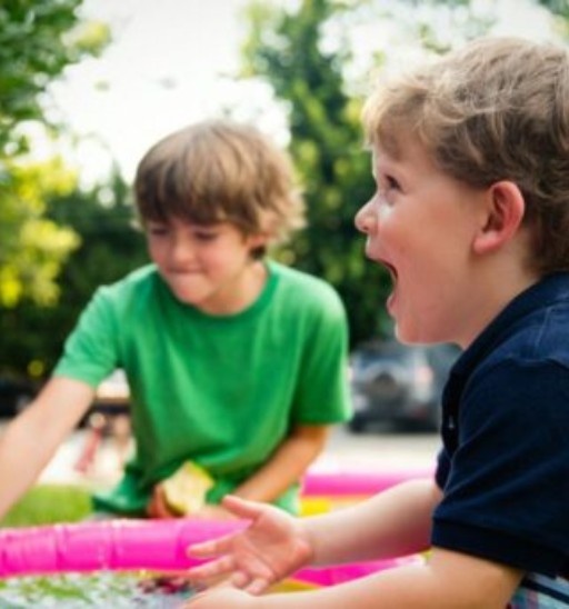 Two young boys playing with water in a backyard. One boy, in a black shirt, is laughing energetically. The other, in green, appears focused. Sunny day.